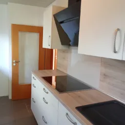 Nowoczesna kuchnia z białymi szafkami, drewnianym blatem i czarnym okapem, widok na płytę indukcyjną i zlew, oraz drzwi z szybą w tle.