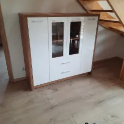Szafa z białymi frontami i drewnianą ramą, z częściowo przeszklonymi drzwiczkami i trzema szufladami, stojąca na drewnianej podłodze, obok drewnianych schodów; na podłodze widoczna skrzynka...