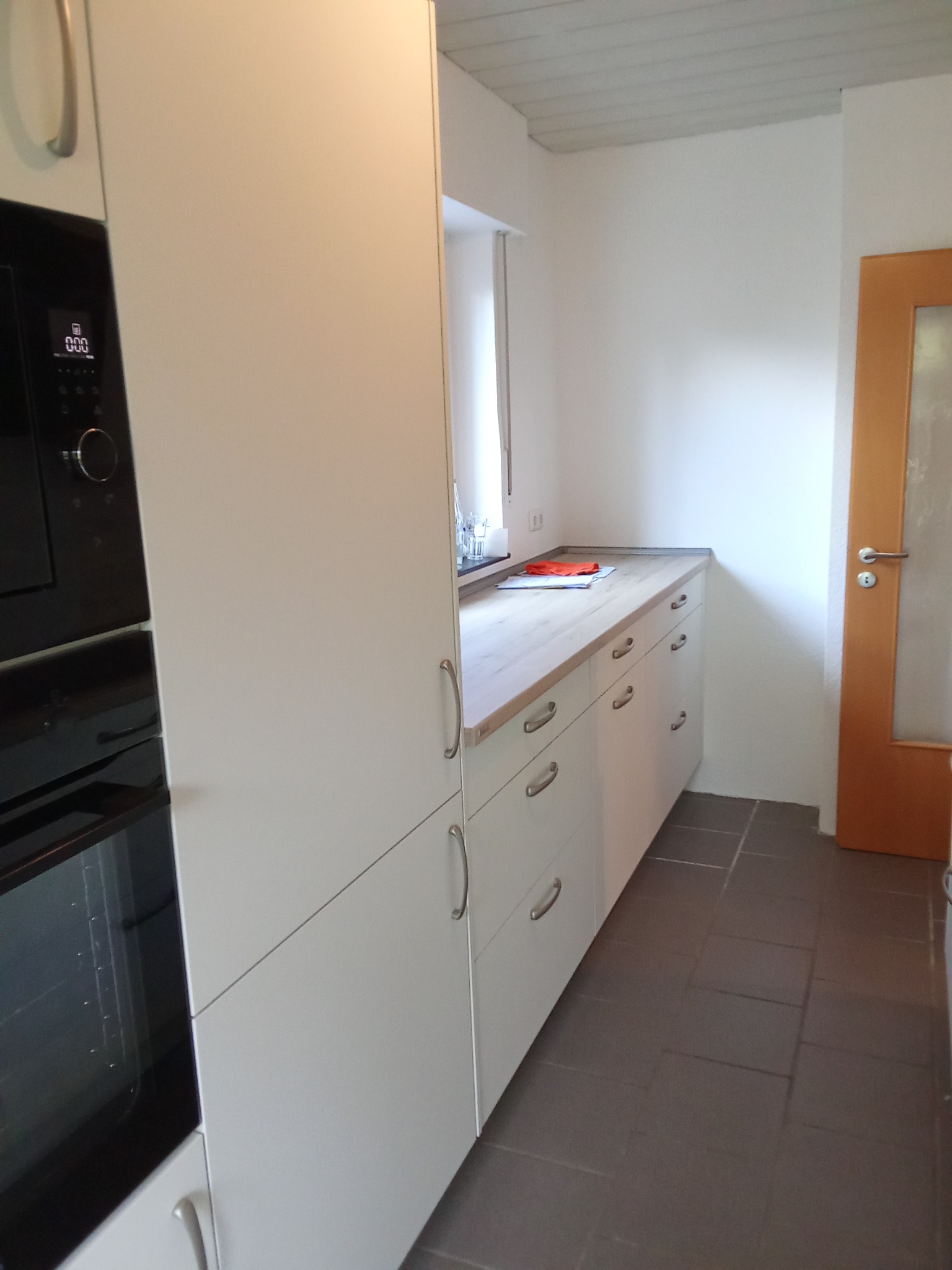 Nowoczesna, biała zabudowa kuchenna z piekarnikiem w zabudowie, szufladami z srebrnymi uchwytami i jasnym blatem, widok z boku na tle białych ścian i szarej podłogi.