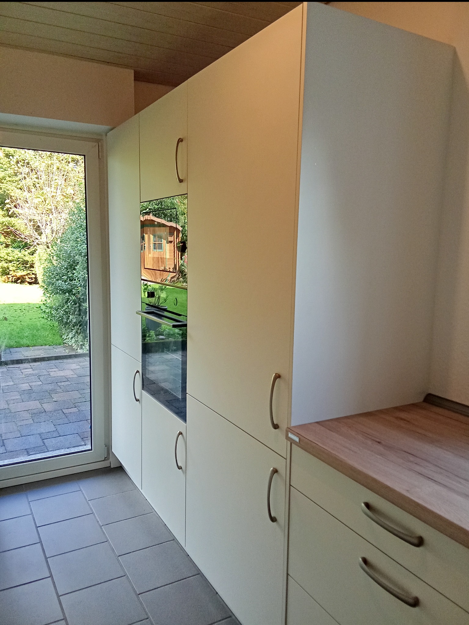 Biała zabudowa kuchenna z piekarnikiem w nowoczesnym stylu, widok z boku, z blatem drewnianym i szarymi płytkami podłogowymi, przez okno widoczny ogród z domkiem.