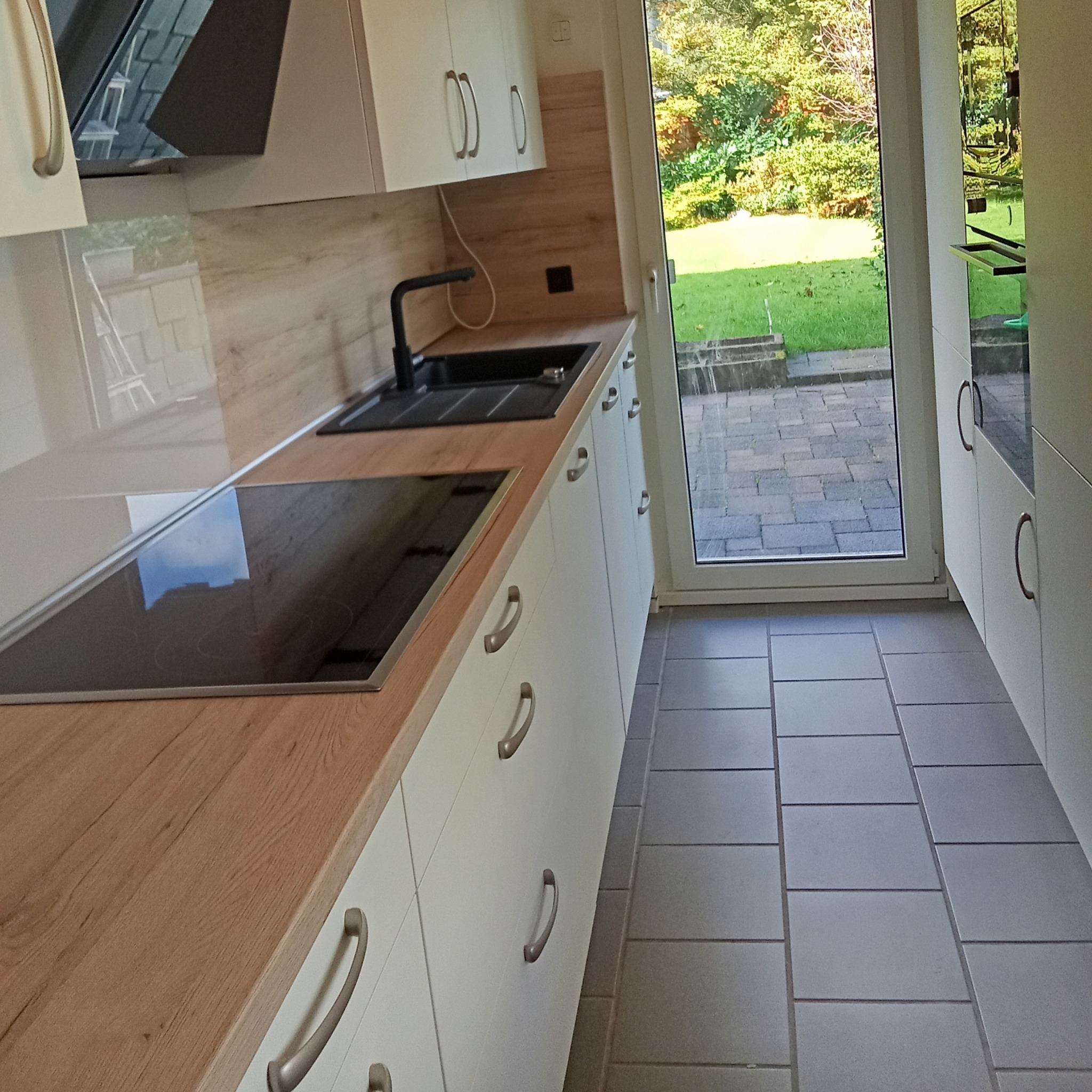 Nowoczesna kuchnia z białymi szafkami, drewnianym blatem, indukcyjną płytą grzewczą i czarnym zlewem, widok na ogród przez przeszklone drzwi.