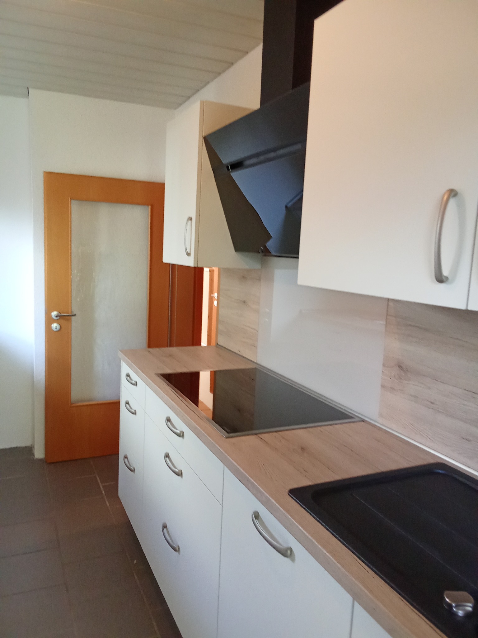 Nowoczesna kuchnia z białymi szafkami, drewnianym blatem i czarnym okapem, widok na płytę indukcyjną i zlew, oraz drzwi z szybą w tle.