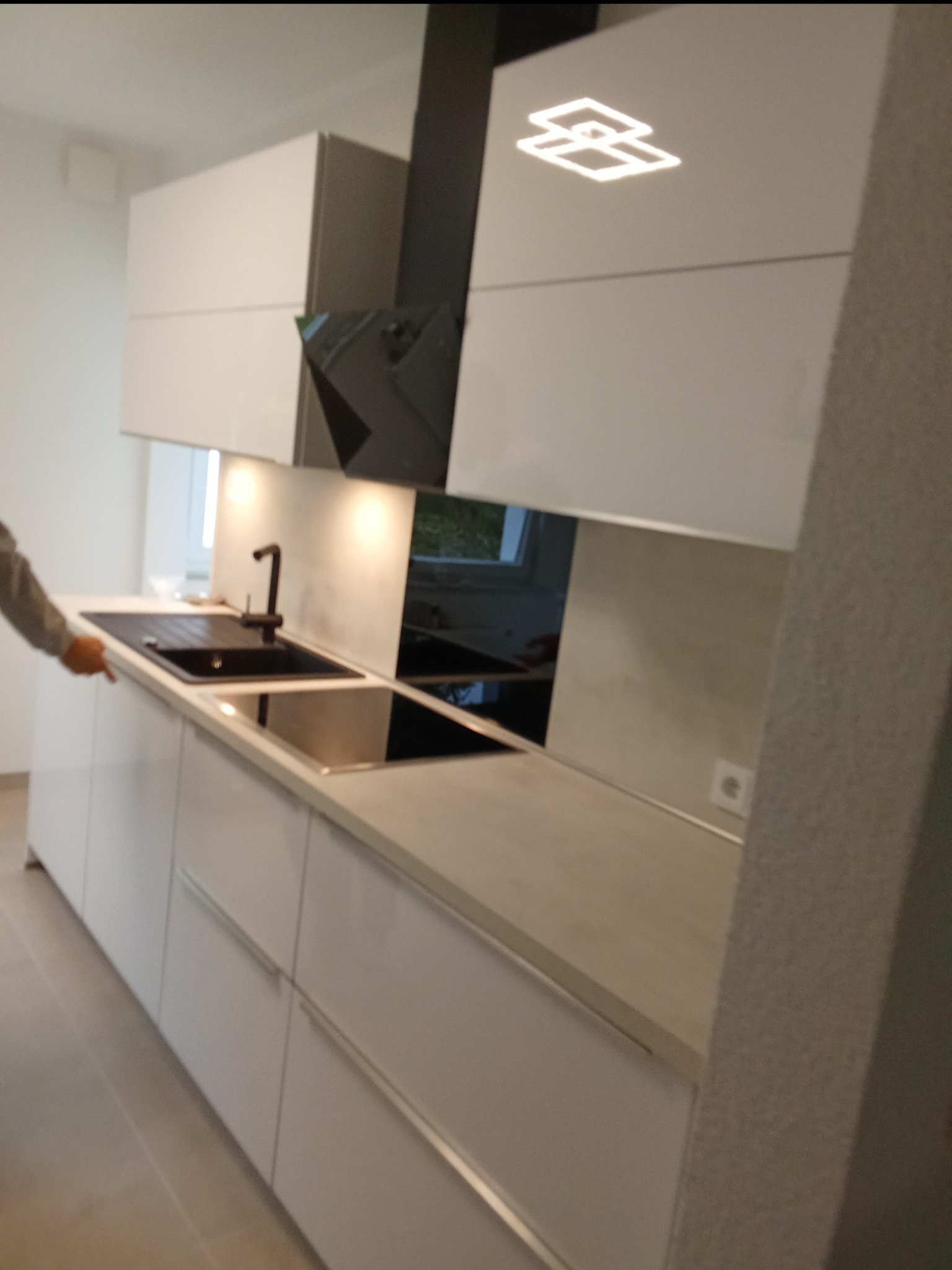 Nowoczesna, biała kuchnia z czarnym zlewem i płytą indukcyjną, minimalistyczny okap z designerskim oświetleniem, widoczny fragment osoby wskazującej na zlew.