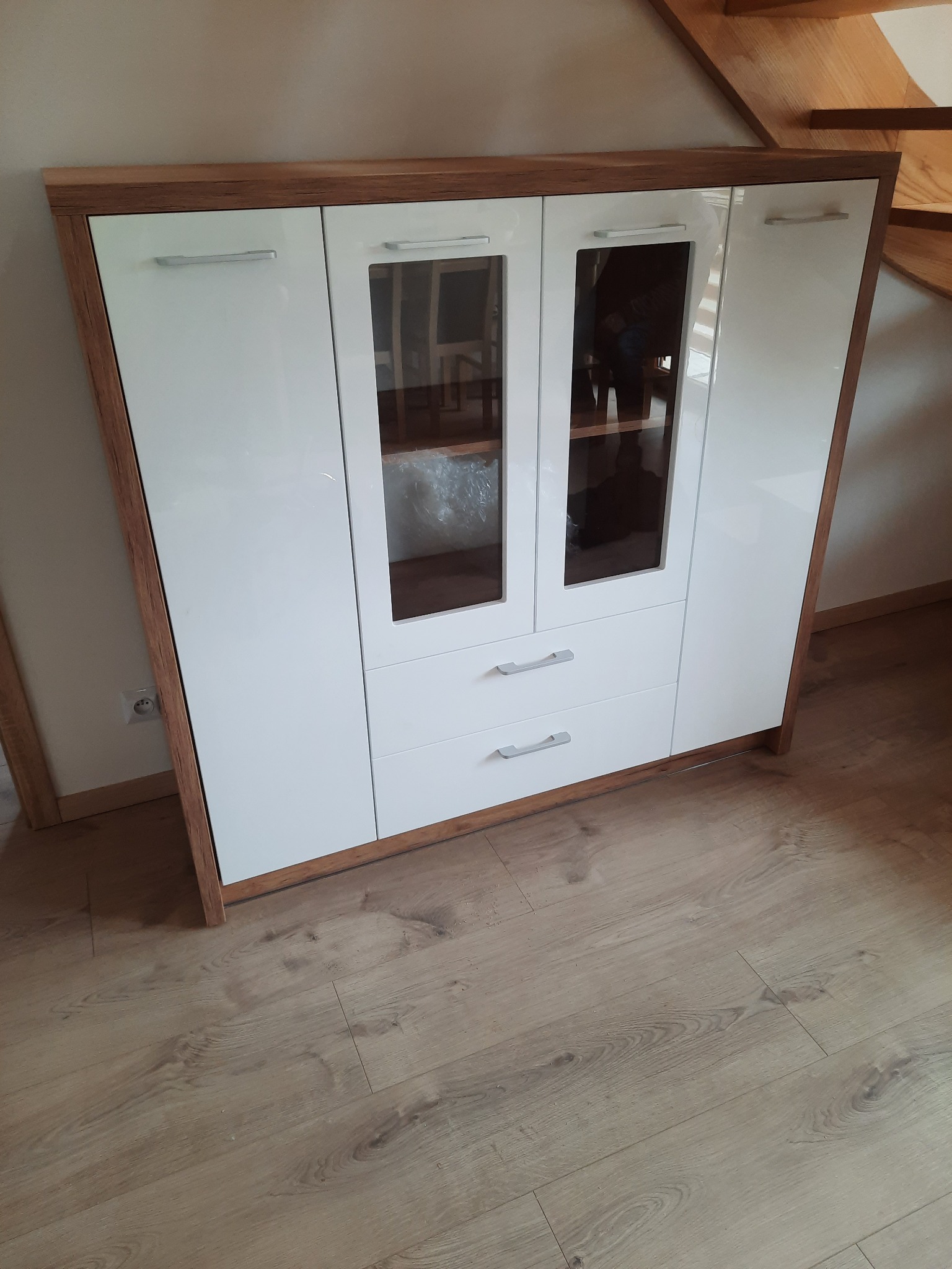 Biała szafa z przeszklonymi drzwiczkami i drewnianą ramą, widoczna na tle drewnianych schodów i podłogi z paneli.