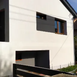 Nowoczesny dom z białą elewacją, szarą stolarką okienną i czarnym dachem, widziany zza grafitowego ogrodzenia z betonowym słupkiem.