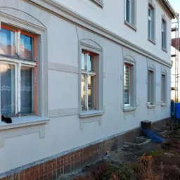 Odnowiona elewacja budynku z oknami w pomarańczowych ramach, widoczne ślady po myciu na cokole i rusztowanie w tle.