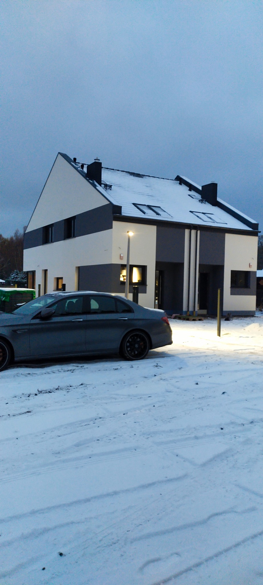 Nowoczesny dom dwukondygnacyjny z płaskim dachem, ocieplony i pomalowany w odcieniach bieli i szarości, częściowo pokryty śniegiem, z szarym samochodem na pierwszym planie, sfotografowany...