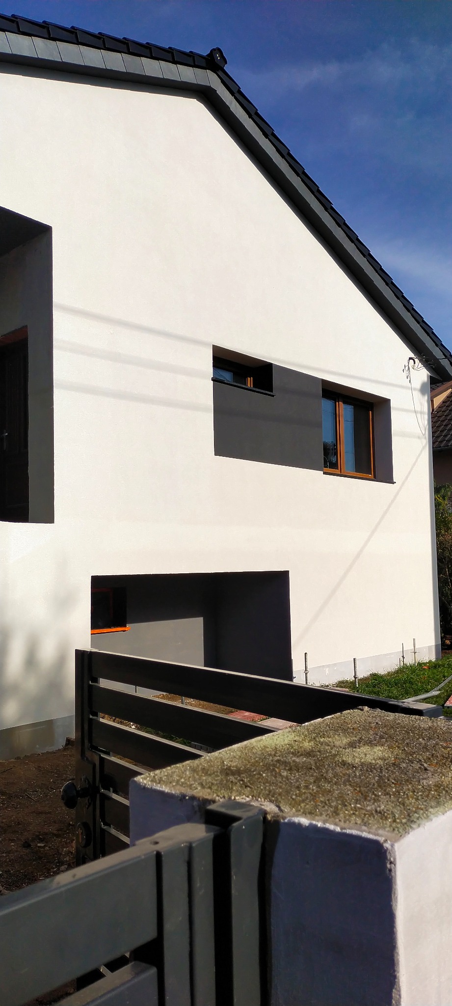 Nowoczesny dom z białą elewacją, szarą stolarką okienną i czarnym dachem, widziany zza grafitowego ogrodzenia z betonowym słupkiem.