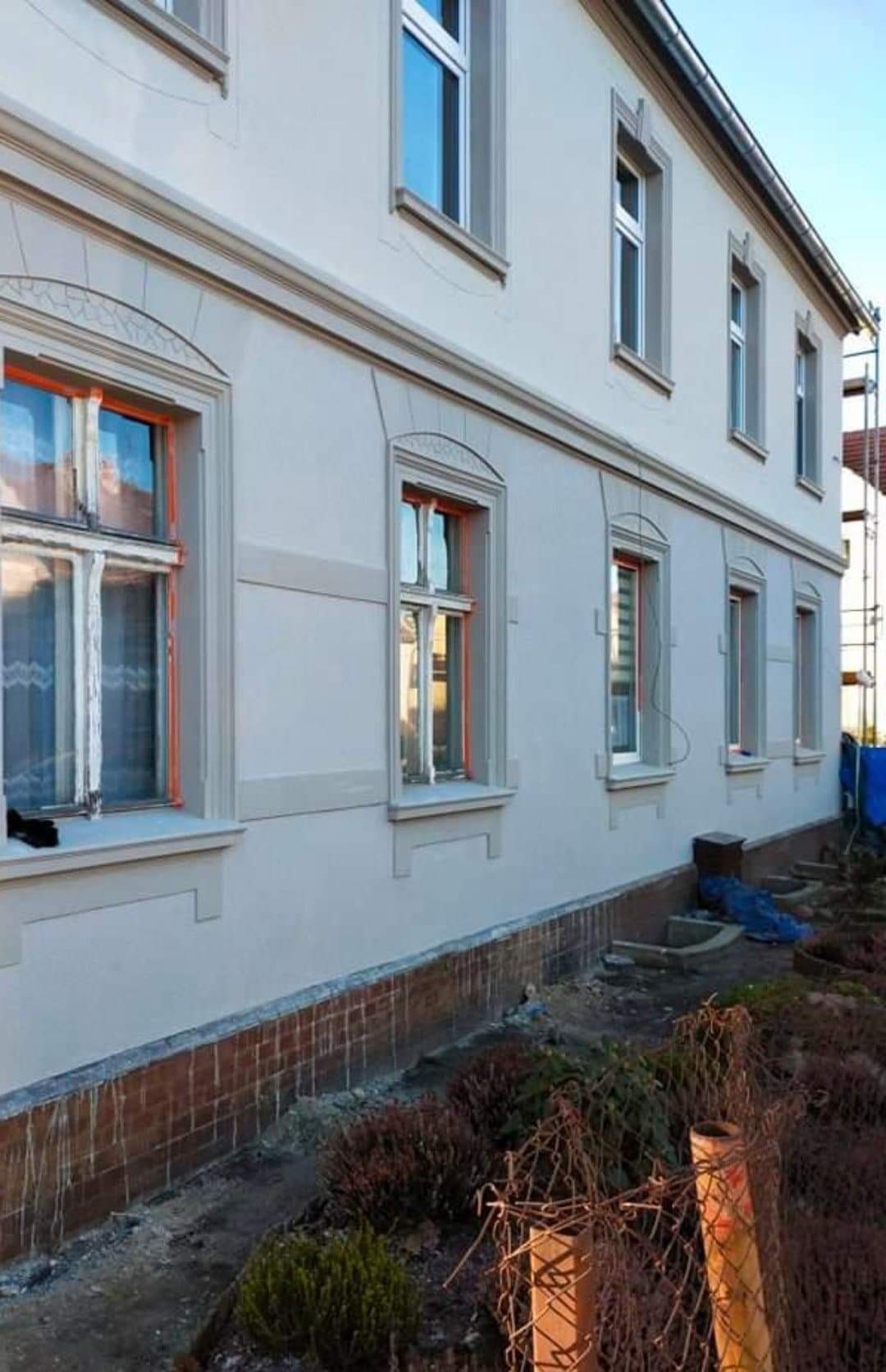 Odnowiona elewacja budynku z oknami w pomarańczowych ramach, widoczne ślady po myciu na cokole i rusztowanie w tle.