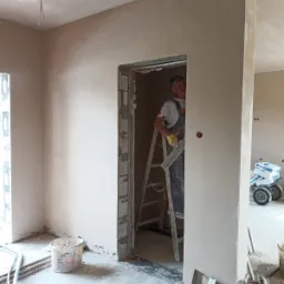 Pracownik w kombinezonie na drabinie, w trakcie nakładania gładzi na ścianę w nowym budynku, widoczne worki z zaprawą na wózku.