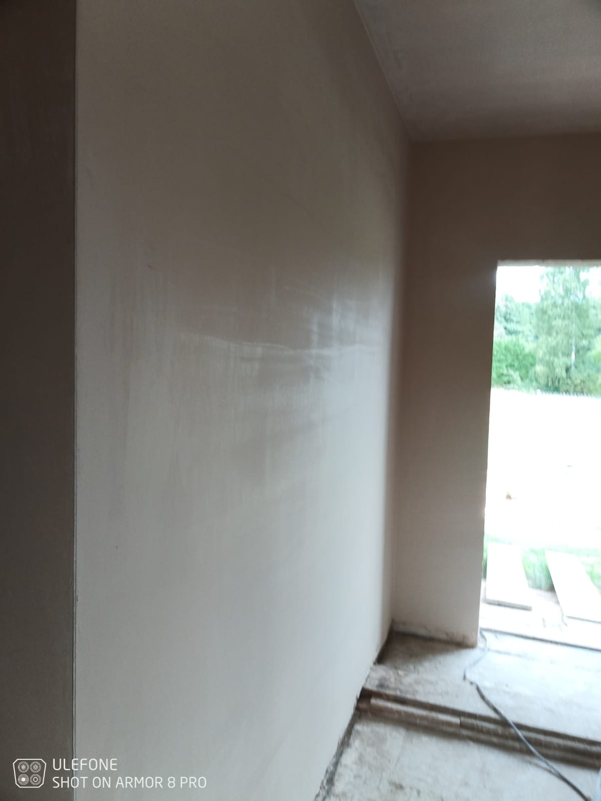 Świeżo nałożony, gładki tynk na ścianie wewnętrznej, widoczne delikatne smugi i przejścia tonalne, z otwartymi drzwiami w tle i widokiem na zieleń na zewnątrz.