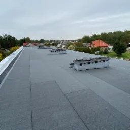 Szary dach pokryty papą termozgrzewalną z kominkami wentylacyjnymi na tle zabudowy jednorodzinnej i pochmurnego nieba.