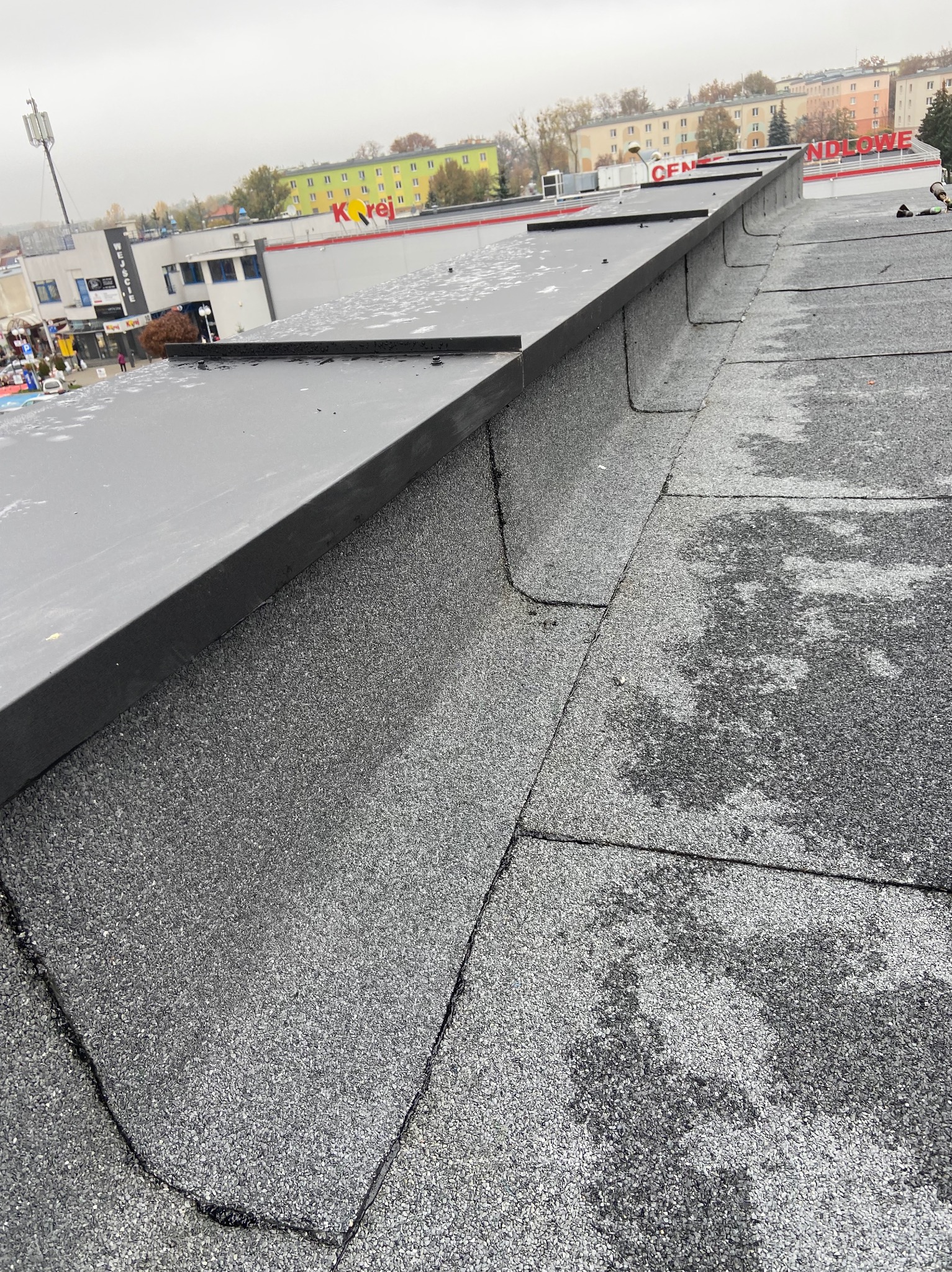 Widok z góry na fragment dachu pokrytego papą, z zamontowaną obróbką blacharską przy krawędzi, na tle budynków miejskich w pochmurny dzień.