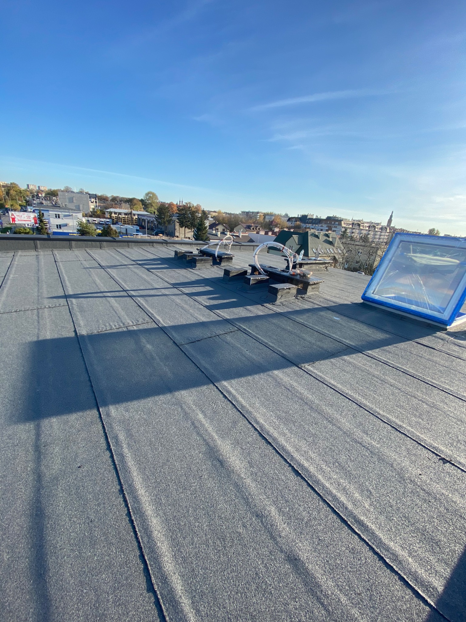 Widok na płaski dach pokryty papą, z widocznymi elementami wentylacji i nowo zamontowanym oknem dachowym z niebieską ramą, na tle zabudowy miejskiej i błękitnego nieba.
