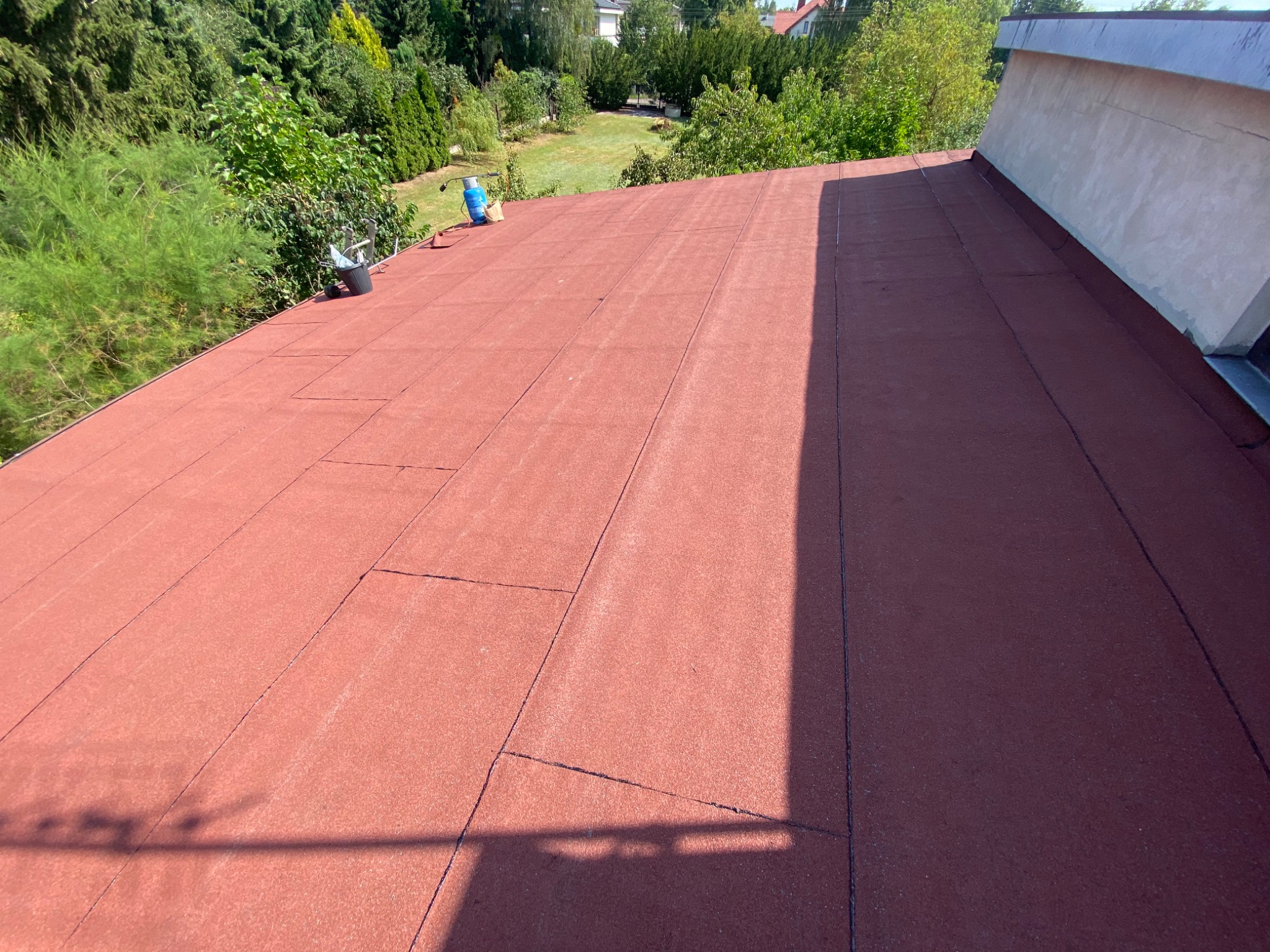 Czerwony, płaski dach pokryty papą termozgrzewalną z widocznymi łączeniami, na tle zieleni drzew i budynków mieszkalnych.
