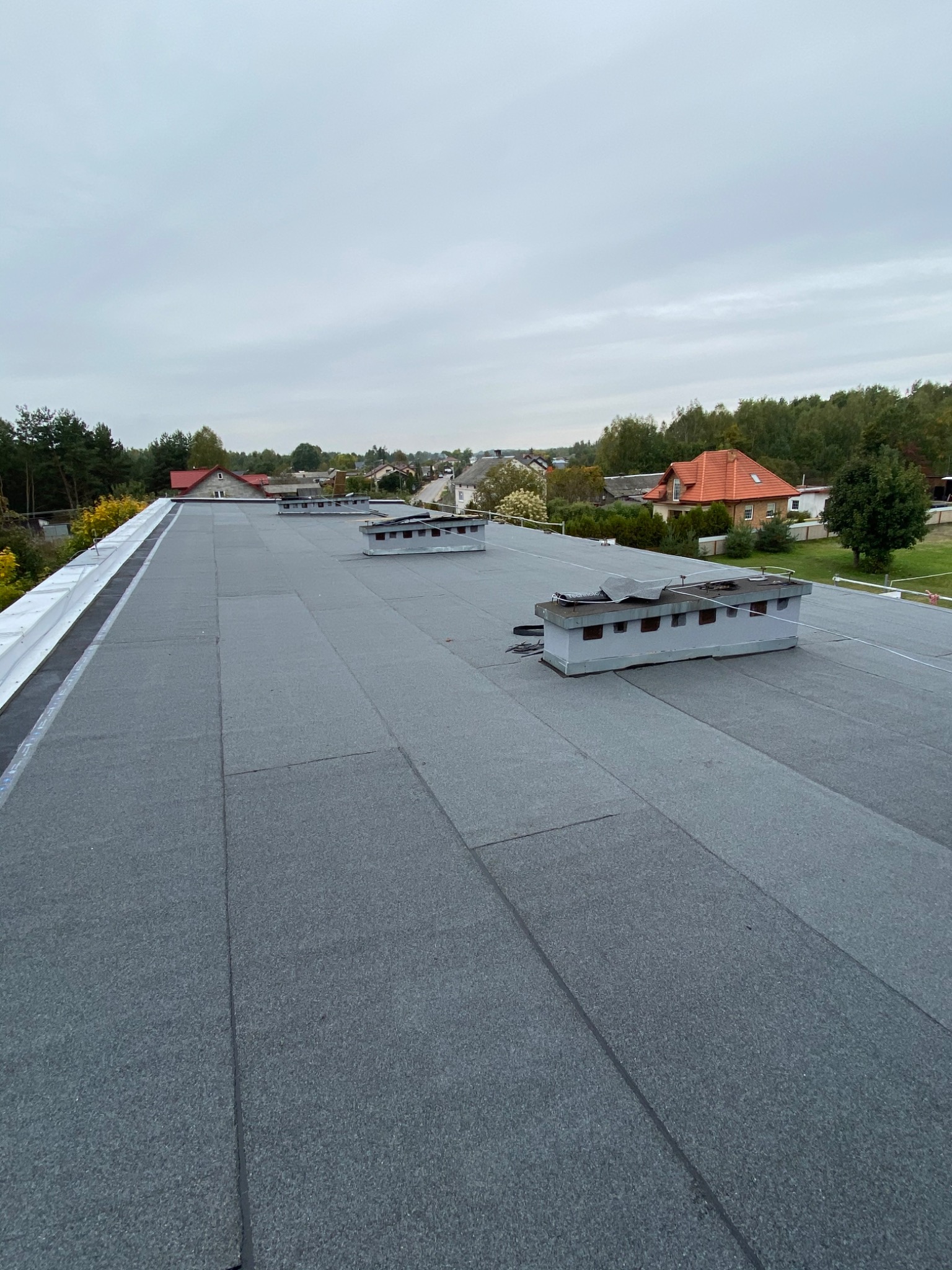 Szary dach pokryty papą termozgrzewalną z kominkami wentylacyjnymi na tle zabudowy jednorodzinnej i pochmurnego nieba.