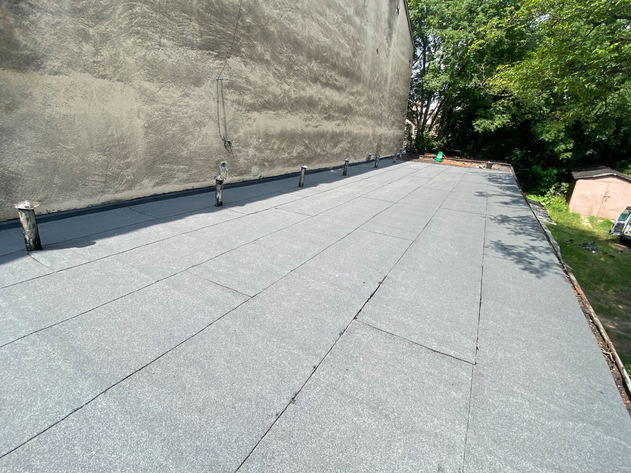 Szary, płaski dach pokryty papą termozgrzewalną, z widocznymi kominkami odpowietrzającymi i ścianą budynku w tle.