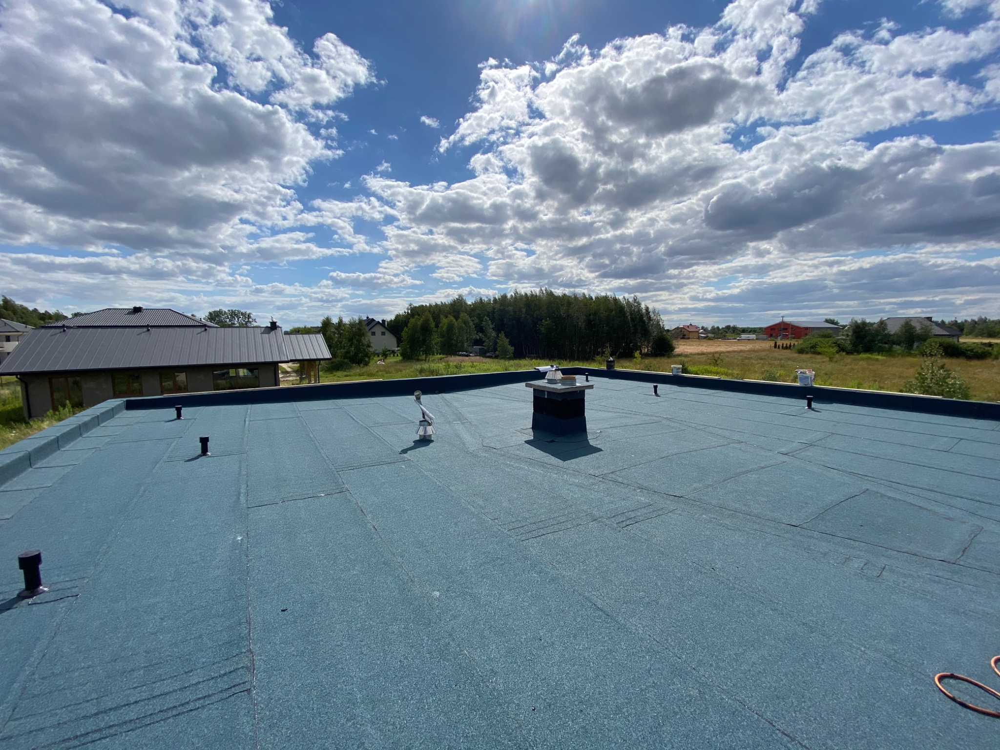 Widok z góry na płaski dach pokryty niebieską papą termozgrzewalną, z kominem i odpowietrznikami na tle błękitnego nieba z chmurami i zielenią w oddali.
