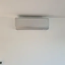 Nowoczesny, srebrny klimatyzator ścienny zamontowany w jasnym pomieszczeniu z widokiem na zewnątrz przez okno, z widocznym panelem sterowania na ścianie.