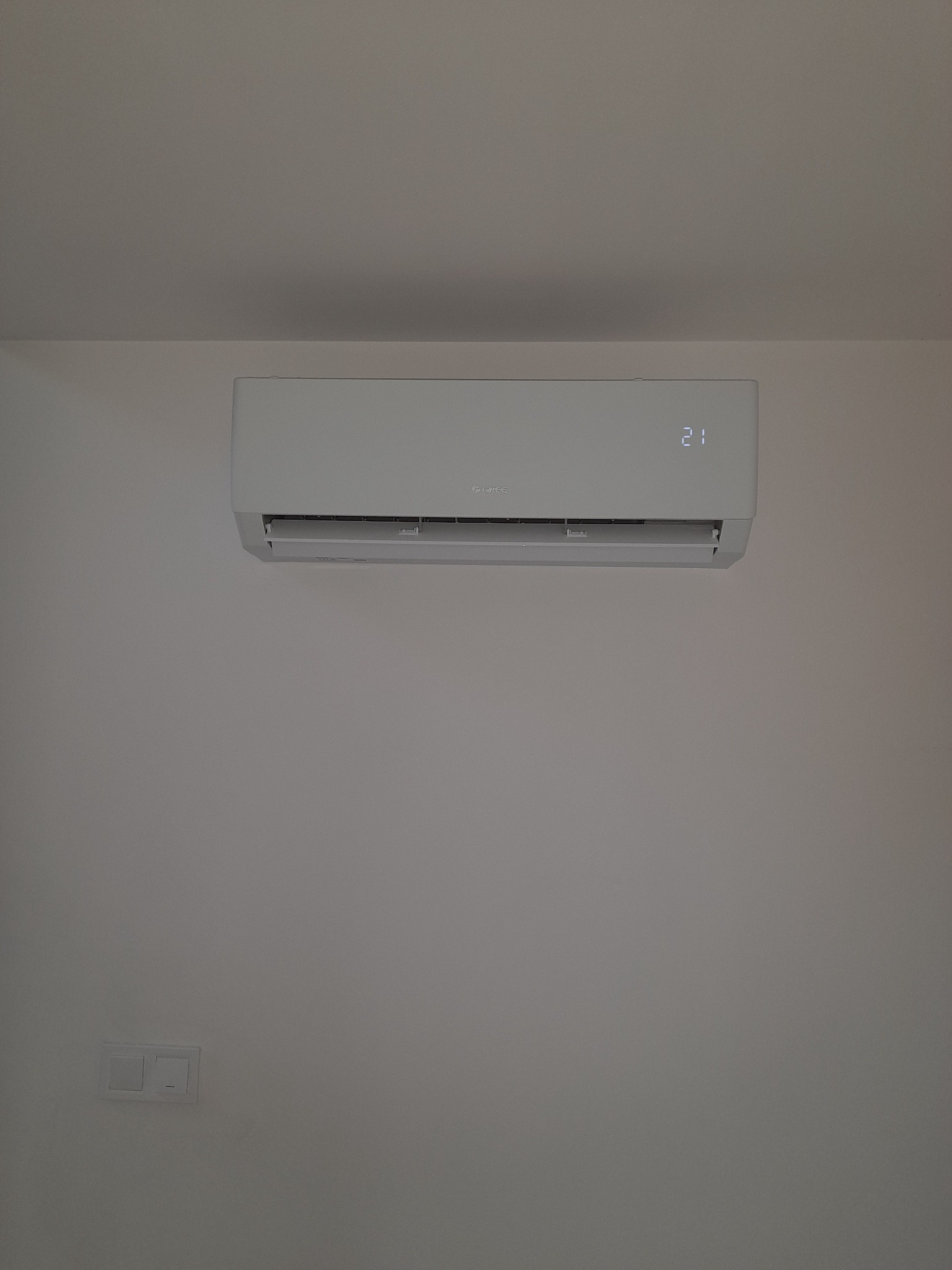 Nowoczesny, biały klimatyzator zamontowany na białej ścianie w pomieszczeniu, wyświetlający temperaturę 21 stopni Celsjusza, widoczny biały włącznik światła na ścianie.