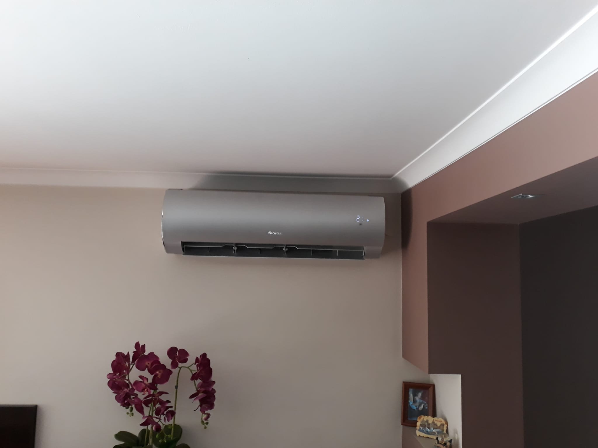 Szary klimatyzator marki Gree zamontowany na beżowej ścianie w pomieszczeniu mieszkalnym, z widocznym wyświetlaczem temperatury i dekoracyjnym bukietem ciemnoróżowych orchidei w tle.