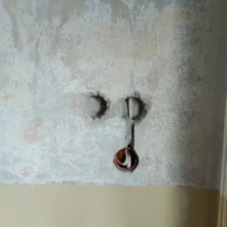 W ścianie z widocznymi śladami po naprawach przygotowane otwory pod instalację elektryczną, z widoczną puszką instalacyjną i przewodami, poniżej grzejnik z leżącymi na nim przedmiotami.