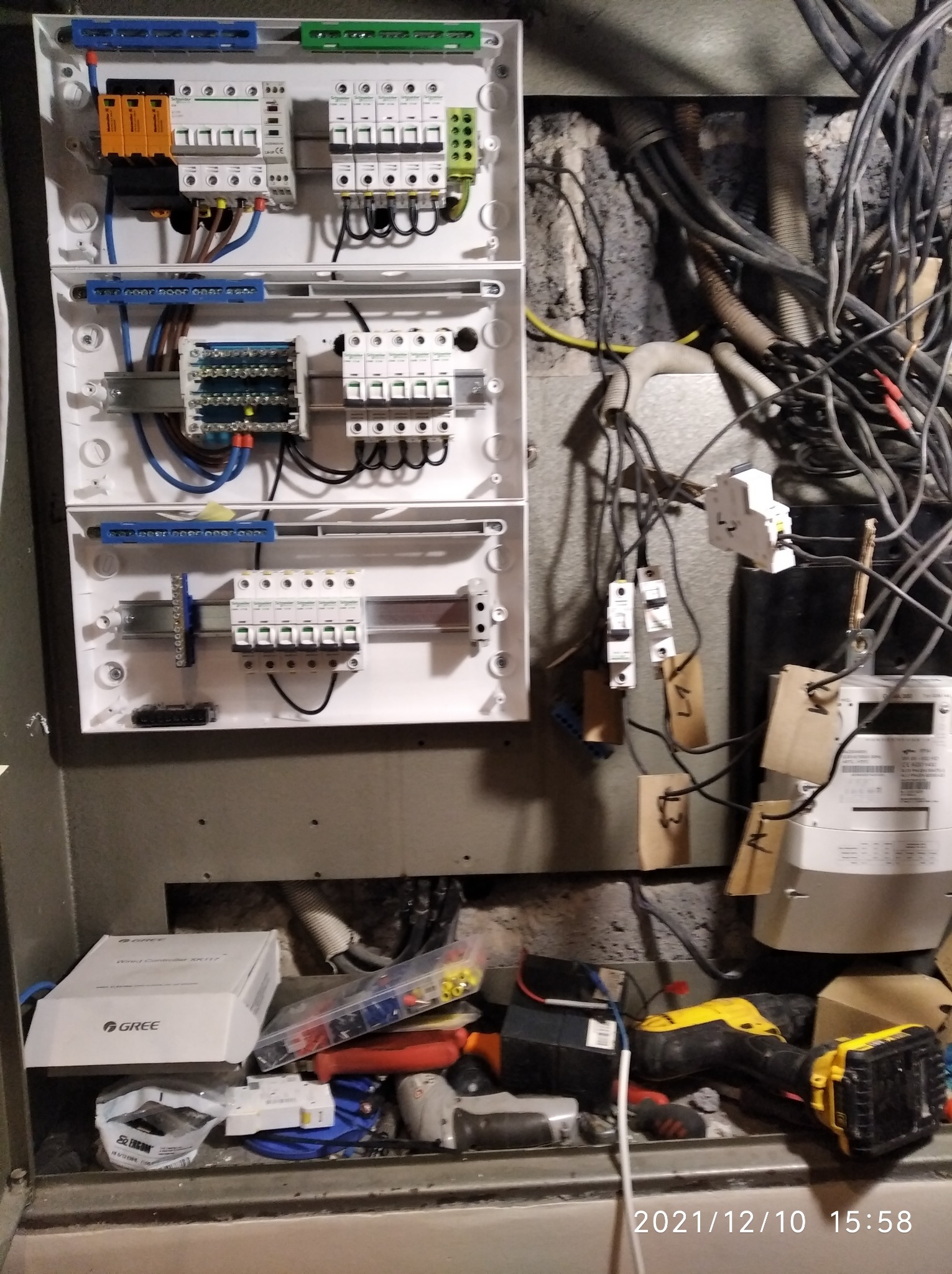 Rozdzielnia elektryczna z widocznymi bezpiecznikami, przewodami i skrzynkami, z narzędziami i elementami instalacji poniżej, widać także licznik energii elektrycznej.