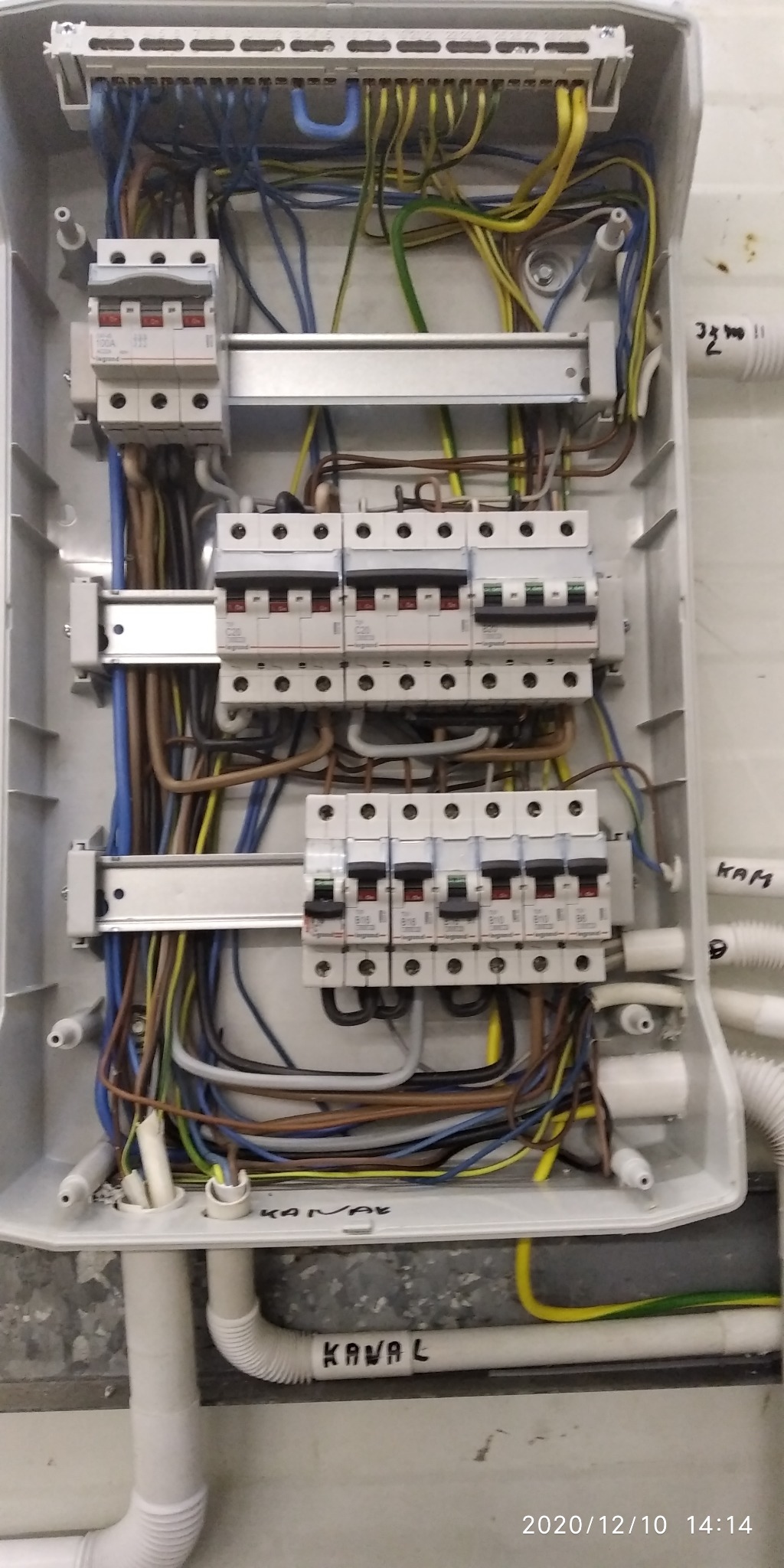 Otwarta skrzynka rozdzielcza z zainstalowanymi bezpiecznikami Legrand i poprowadzonymi przewodami elektrycznymi w różnych kolorach, widoczne napisy na ścianie i rurach instalacyjnych.