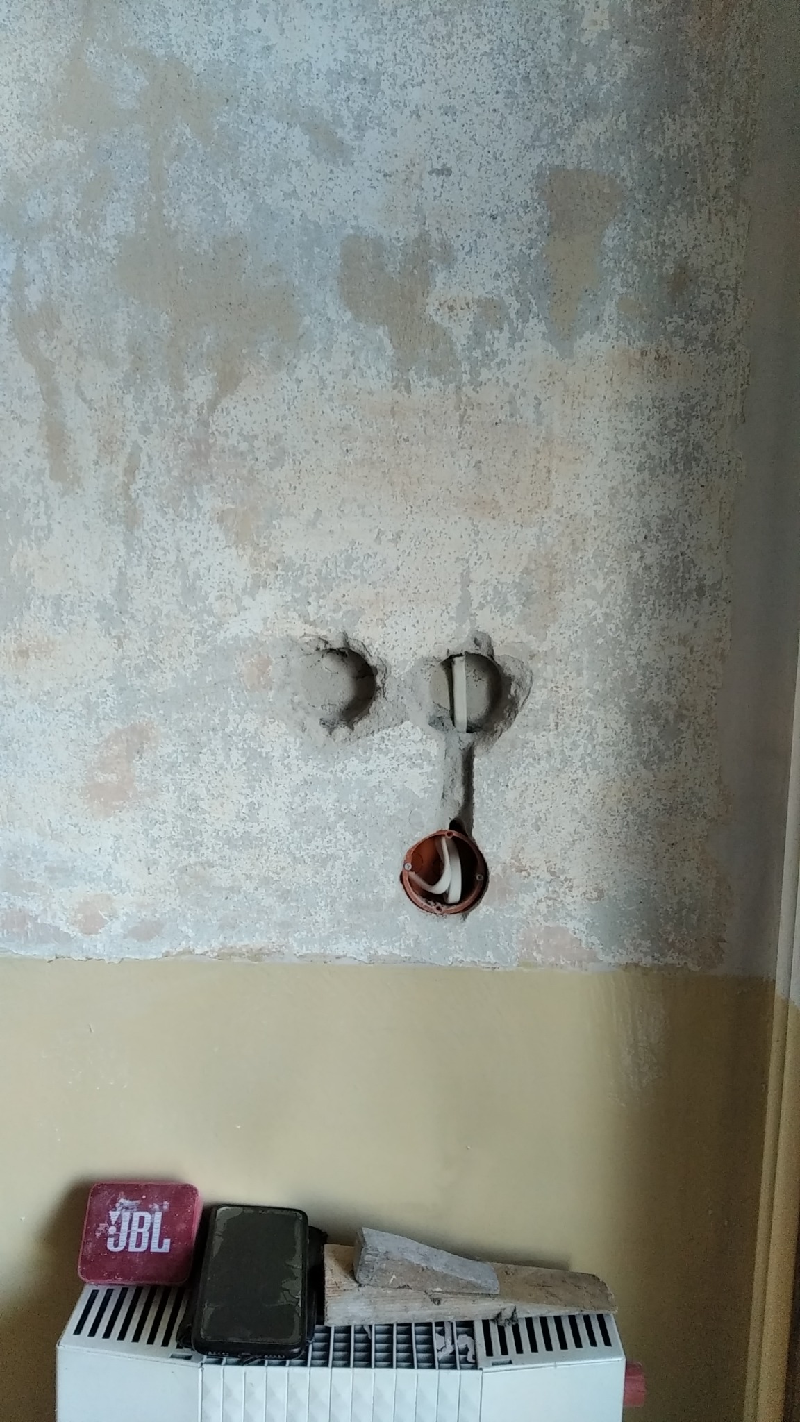 W ścianie z widocznymi śladami po naprawach przygotowane otwory pod instalację elektryczną, z widoczną puszką instalacyjną i przewodami, poniżej grzejnik z leżącymi na nim przedmiotami.