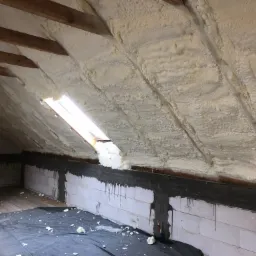 Poddasze z drewnianymi belkami pokryte grubą warstwą białej pianki izolacyjnej, widoczne okno dachowe i fragment muru z cegły.