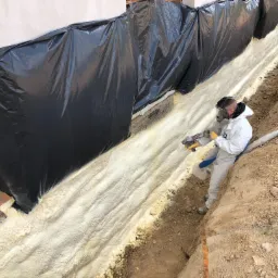 Pracownik w kombinezonie i masce ochronnej natryskuje pianę PUR na zabezpieczoną folią fundamentową ścianę budynku w wykopie.