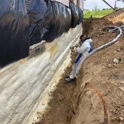 PUR-TECH Paweł Rzęsa - Pracownik w kombinezonie ochronnym natryskuje pianę izolacyjną na fundament budynku, zabezpieczonego czarną folią, widoczne węże doprowadzające materiał, wykop wokół fundamentu.