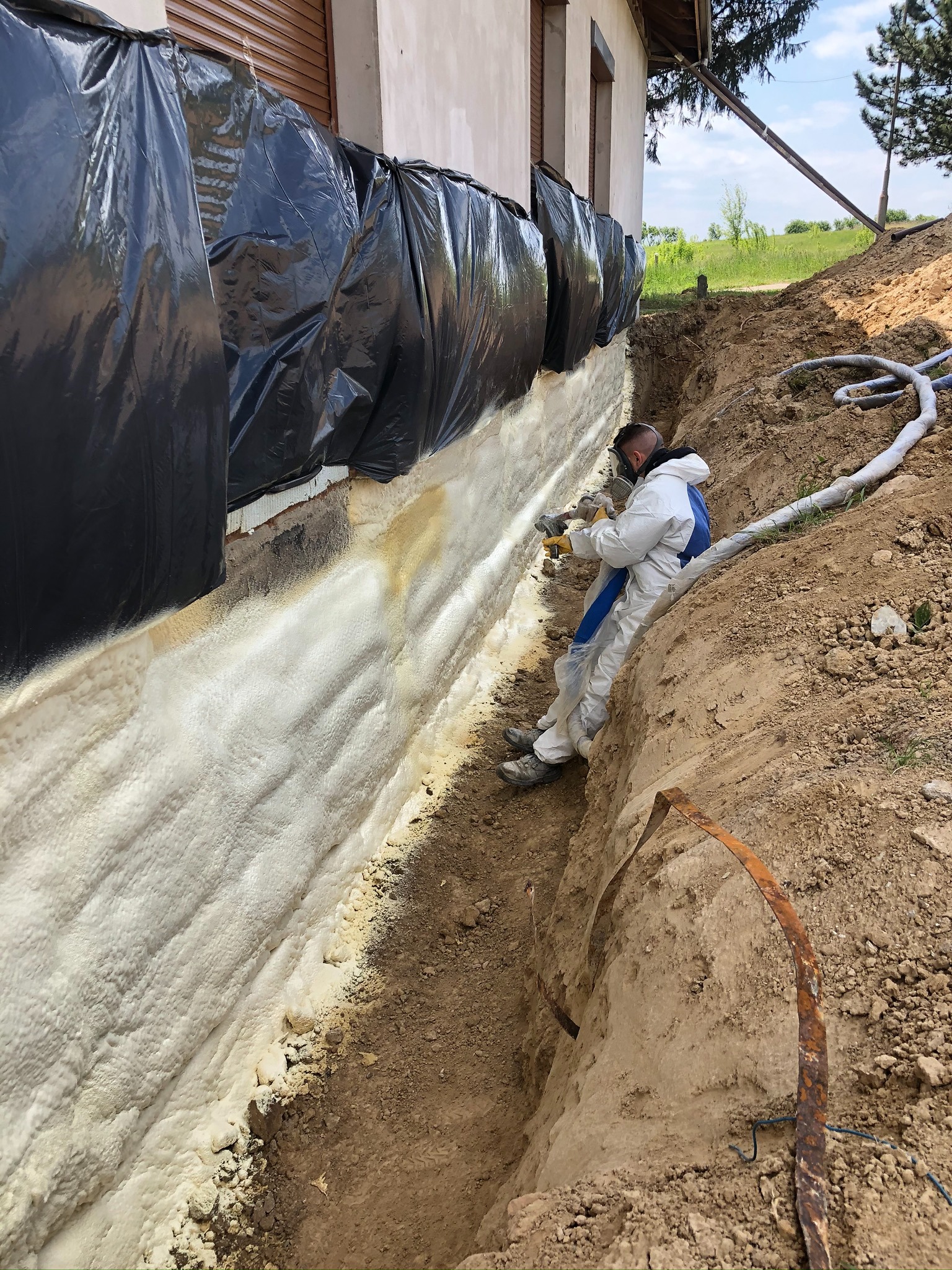 Pracownik w kombinezonie ochronnym natryskuje pianę izolacyjną na fundament budynku, zabezpieczonego czarną folią, widoczne węże doprowadzające materiał, wykop wokół fundamentu.