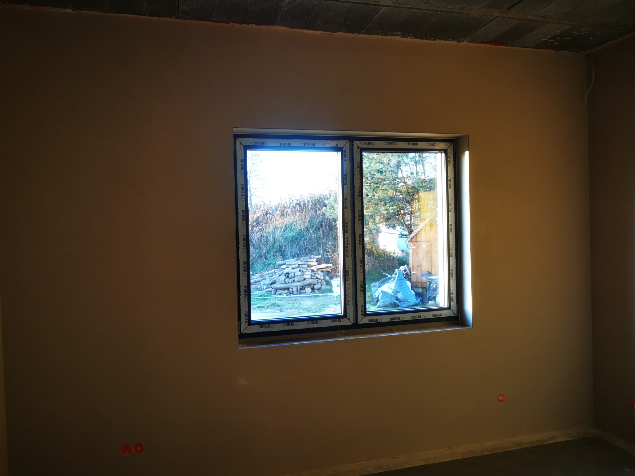 Świeżo nałożona, gładka powierzchnia ścian w pokoju po usłudze w Łodzi, widoczne okno z widokiem na zewnątrz i wystające kable elektryczne.