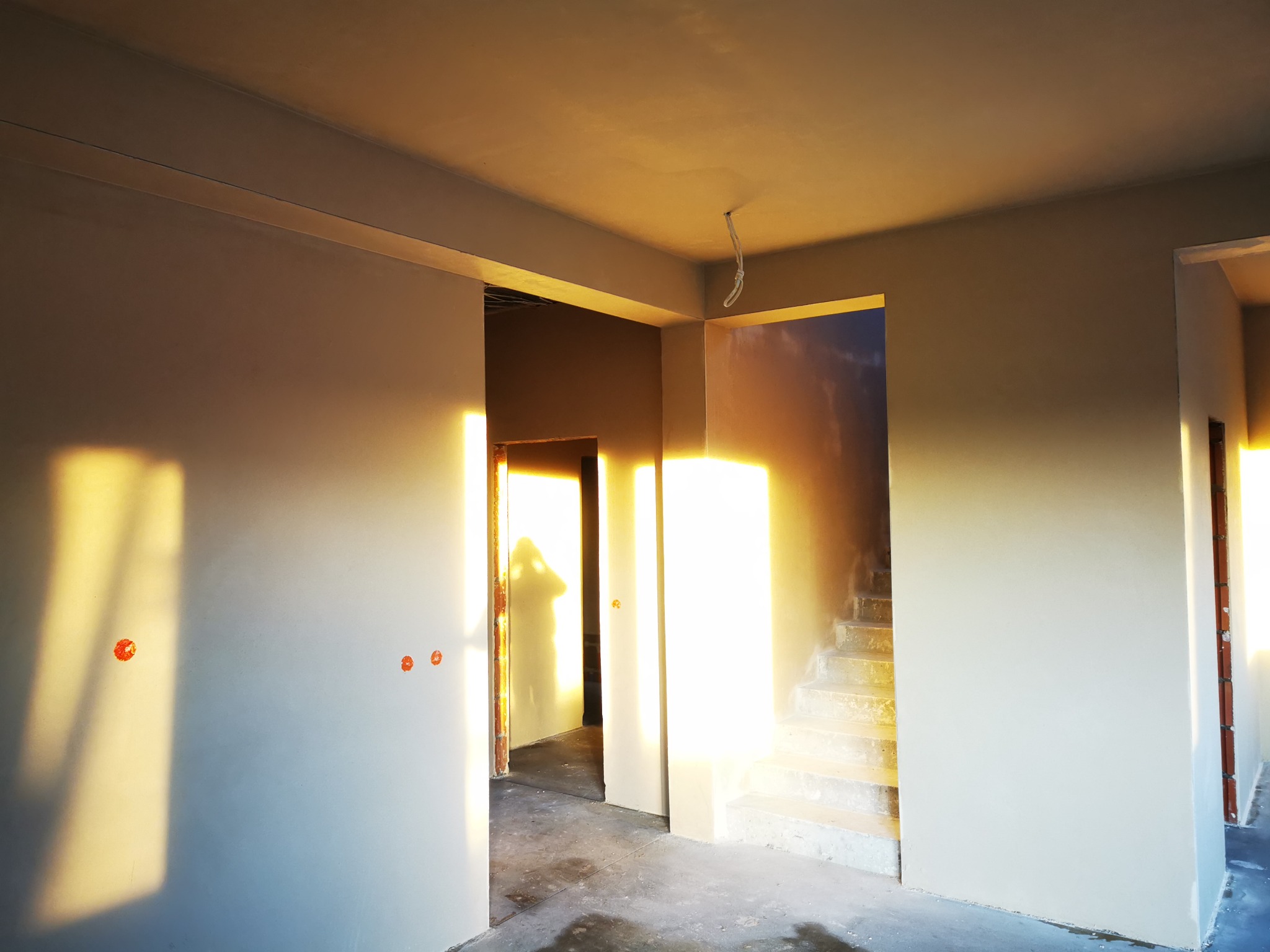 Świeżo otynkowane ściany wewnętrzne w surowym stanie, widoczne ślady po łatach, ciepłe światło słoneczne wpadające przez okna, surowe schody w tle.