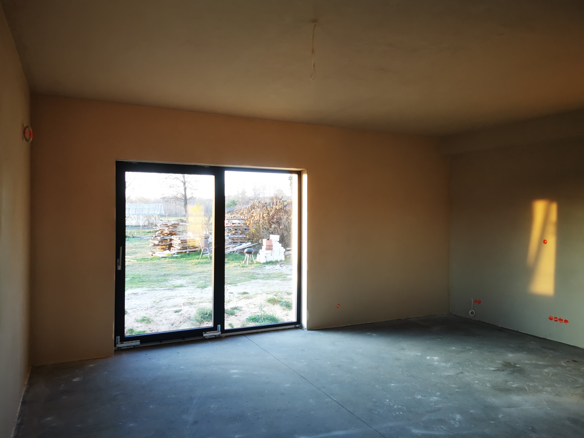 Wnętrze po tynkowaniu maszynowym z widocznymi gładkimi ścianami i oknem balkonowym z widokiem na ułożone palety.