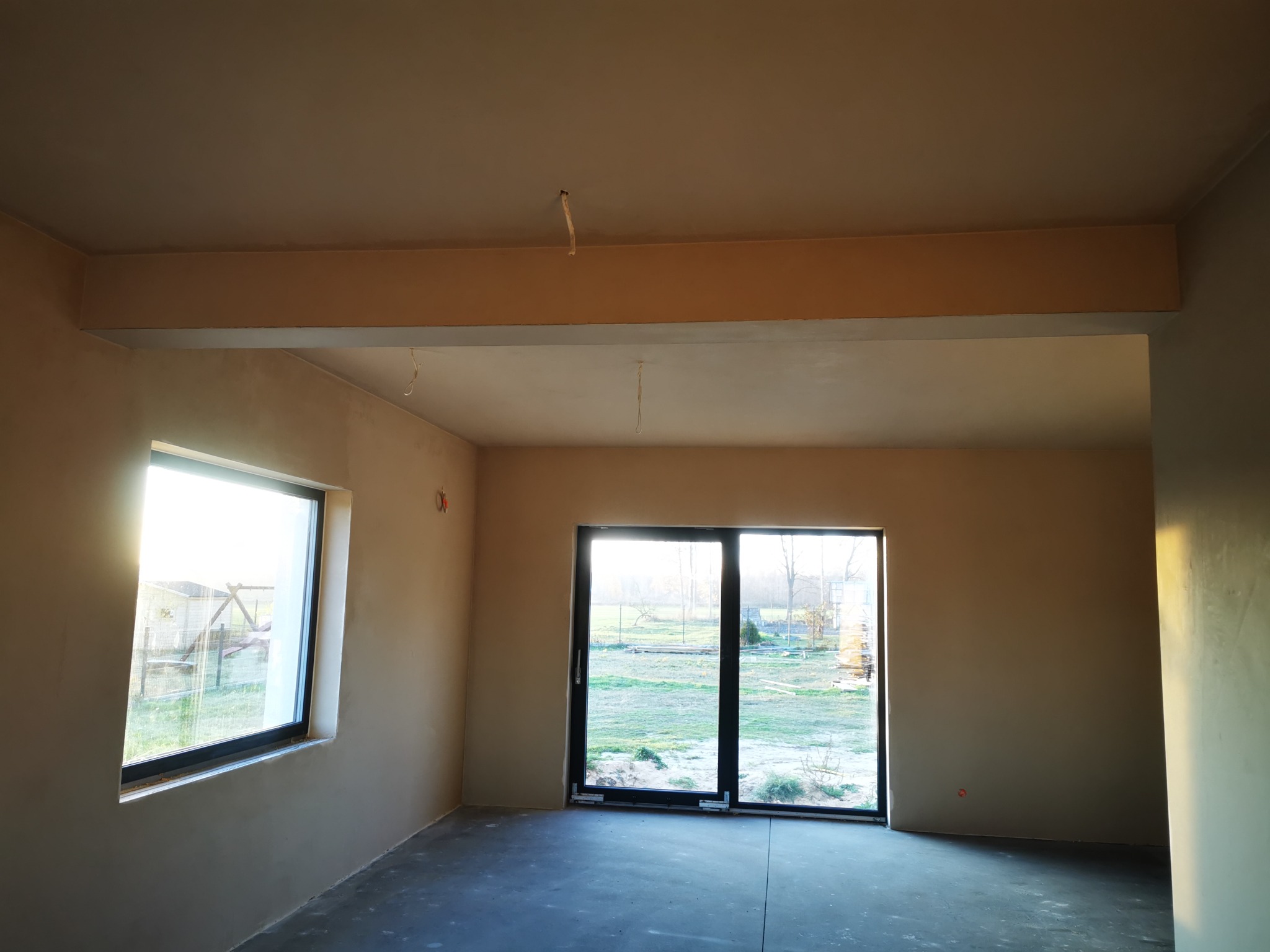 Wnętrze surowego domu z nałożonymi gładziami, widoczne okno z czarną ramą i rozsuwane drzwi balkonowe, przez które widać zieleń na zewnątrz. Na suficie wystają przewody.