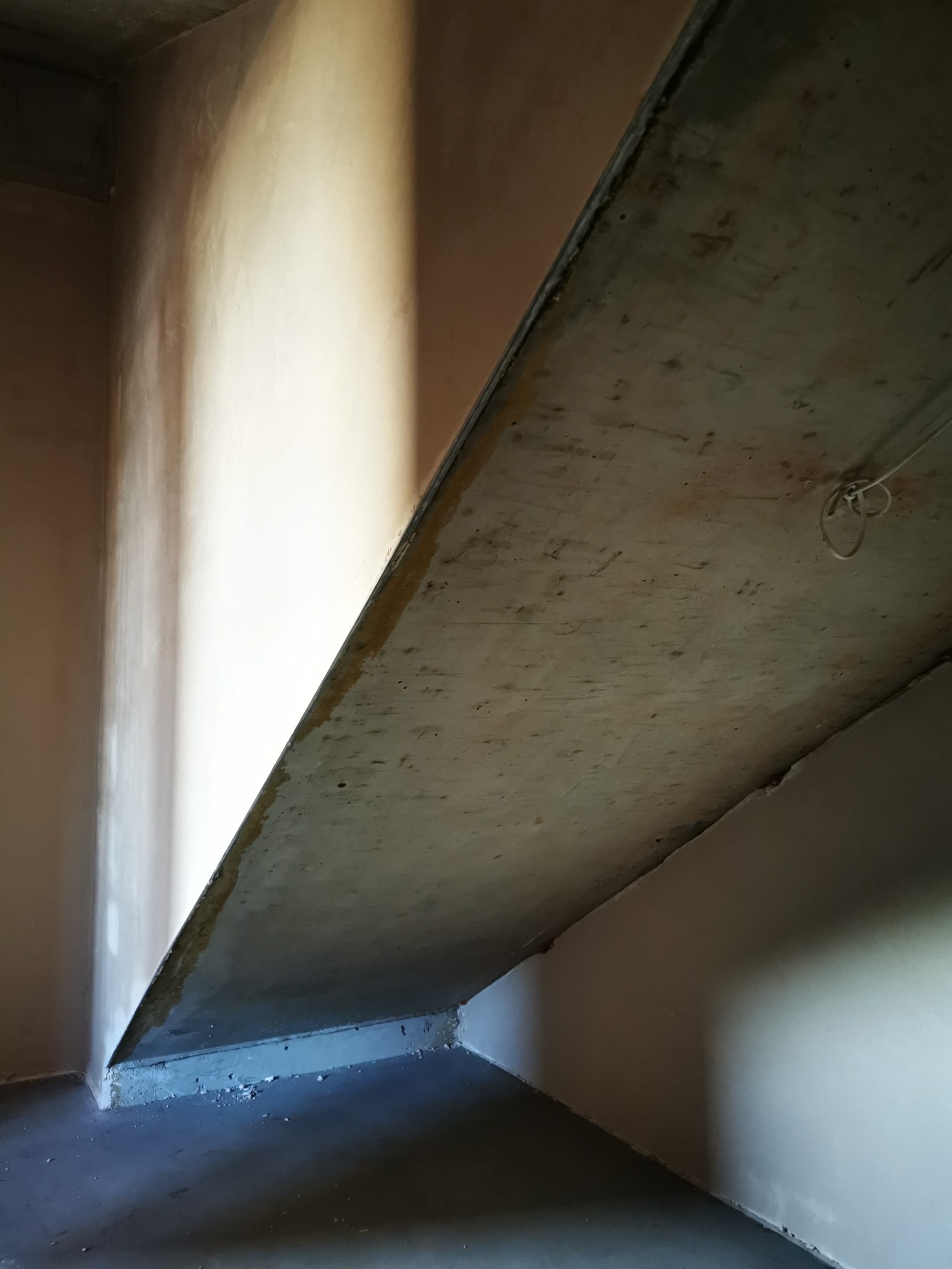 Świeżo położony tynk na ścianie wewnętrznej, widoczny surowy betonowy strop i fragment podłogi, oświetlone naturalnym światłem wpadającym z góry.