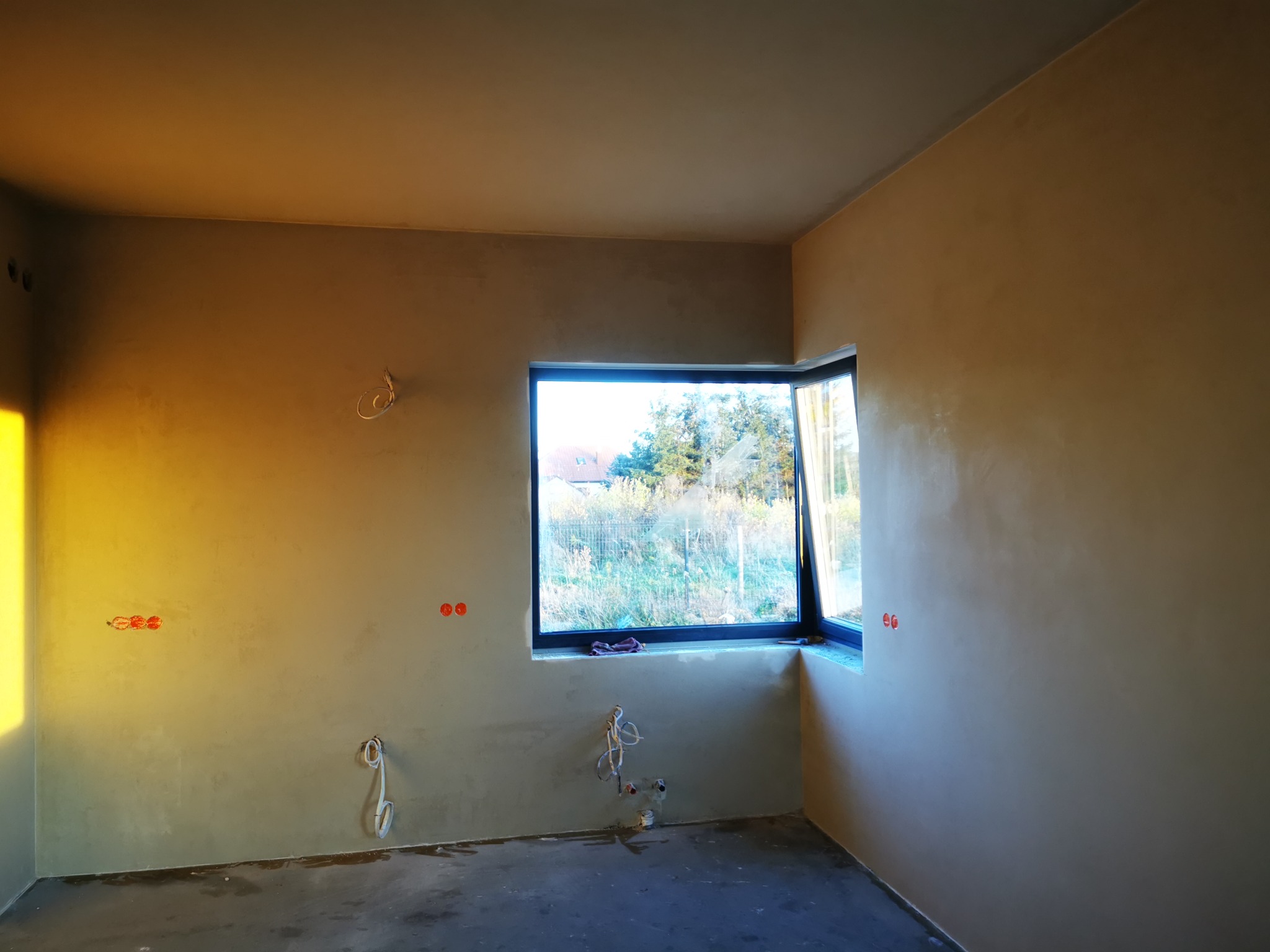 Wnętrze pokoju po nałożeniu świeżych, gładkich tynków; widoczne wystające kable elektryczne i rury instalacyjne przed wykończeniem.