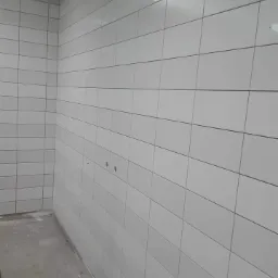 Wnętrze pomieszczenia z białymi, prostokątnymi płytkami ułożonymi na ścianach i suficie, widoczne niedokończone elementy instalacji elektrycznej oraz surowa, betonowa podłoga.