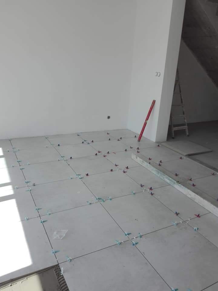 Układanie jasnych płytek podłogowych z użyciem systemu poziomowania, widoczna poziomica i drabina w tle