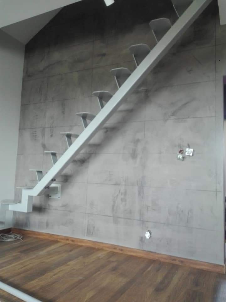 Nowoczesne, minimalistyczne schody wspornikowe w kolorze białym, zamontowane na ścianie wykończonej płytami imitującymi beton, z drewnianą podłogą w tle.