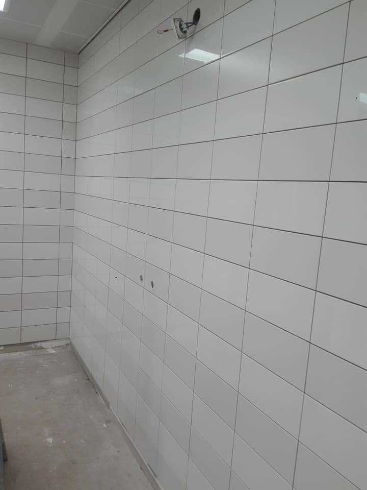 Wnętrze pomieszczenia z białymi, prostokątnymi płytkami ułożonymi na ścianach i suficie, widoczne niedokończone elementy instalacji elektrycznej oraz surowa, betonowa podłoga.