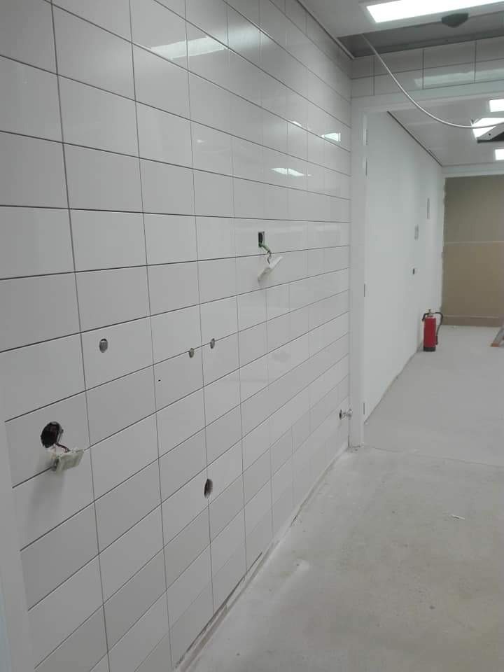 Ściana wyłożona białymi, prostokątnymi płytkami z widocznymi otworami pod gniazdka elektryczne i wystającymi kablami, w tle widoczny korytarz z czerwoną gaśnicą.