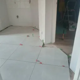 Białe, połyskujące płytki podłogowe w trakcie układania w pomieszczeniu, widoczne czerwone i zielone kliny poziomujące, oraz narzędzia i drabina w tle.