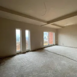 Wnętrze surowego pokoju po nałożeniu tynków, widoczne ślady po maszynowym nakładaniu na podłodze, dwa okna wpuszczające naturalne światło, widoczne kable elektryczne na podłodze i suficie.