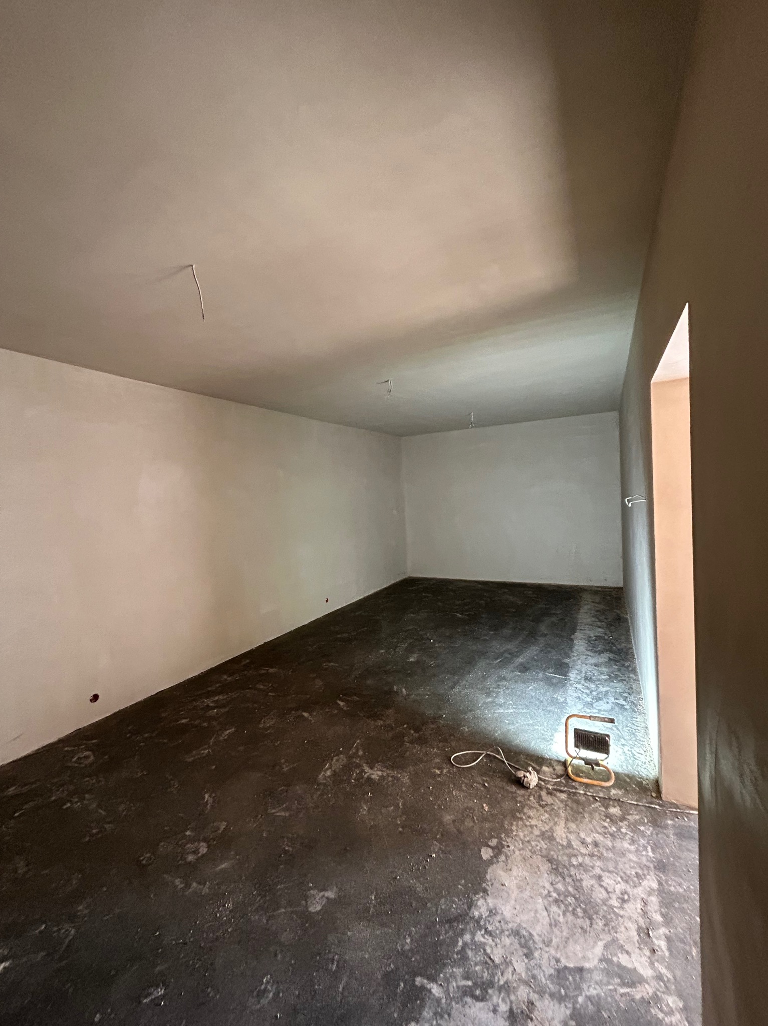 Wnętrze po nałożeniu tynków: gładkie, jasne ściany i sufit, ciemna podłoga. W rogu lampa budowlana oświetla fragment posadzki. Widoczny przewód elektryczny.