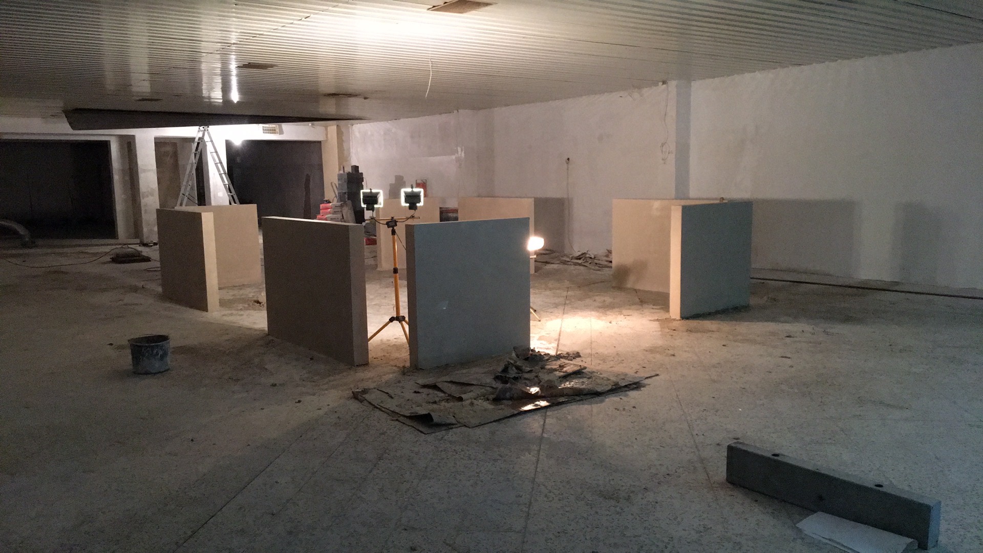 Przygotowanie do tynkowania maszynowego: widoczne płyty gipsowo-kartonowe ustawione pionowo, oświetlenie budowlane na statywach, wiadro i resztki materiałów na surowym betonowym podłożu w dużym...