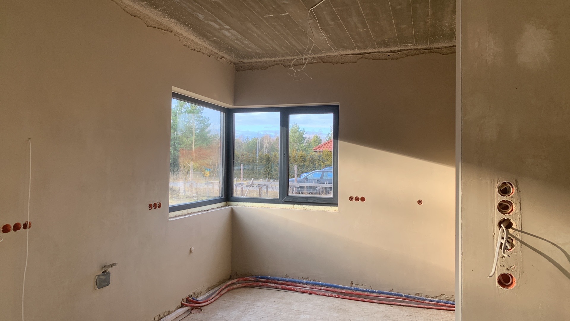 Wnętrze w stanie deweloperskim po nałożeniu tynków, widoczne instalacje elektryczne i hydrauliczne, okno narożne z widokiem na zewnątrz, surowy betonowy strop.