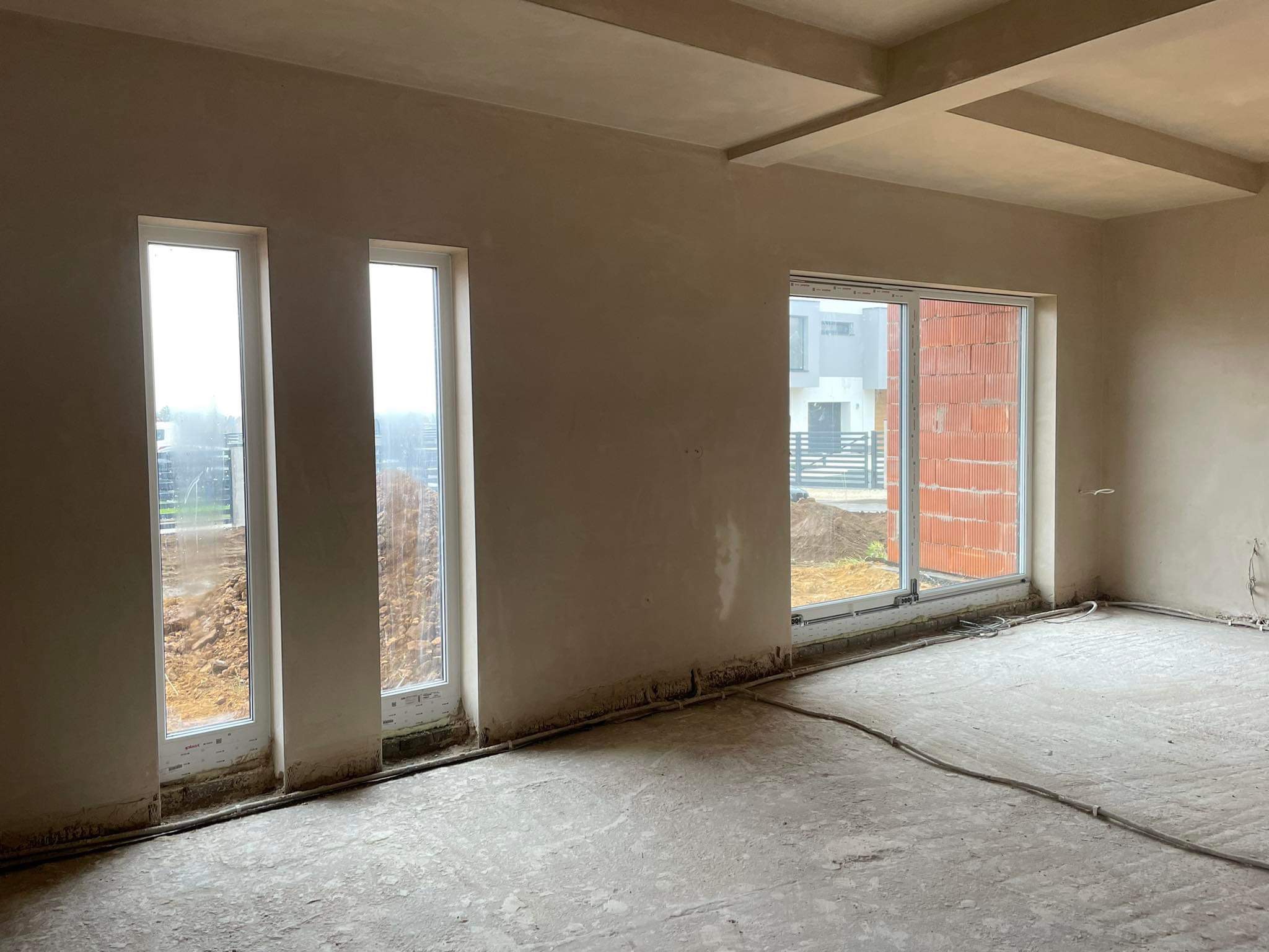 Wnętrze surowego domu po nałożeniu gładzi gipsowej na ściany i sufit, widoczne okna z widokiem na zewnątrz oraz wystające kable elektryczne.